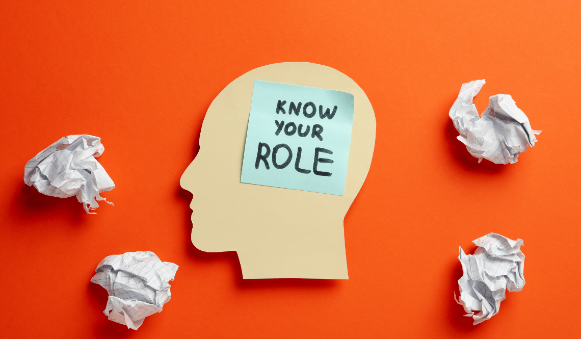Sagoma di testa umana su sfondo arancione con un post-it azzurro sulla fronte che riporta la scritta “Know your role”, circondata da fogli accartocciati.