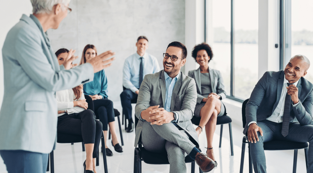 Gruppo di professionisti in abbigliamento formale seduti in una sala luminosa, mentre una persona più anziana conduce una sessione formativa.