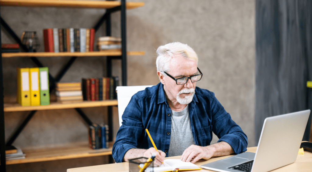 Un uomo maturo in abbigliamento casual elegante e con gli occhiali sta usando un computer portatile per studiare online da casa/ufficio. Un uomo dai capelli grigi sta scrivendo su un quaderno mentre è seduto e guarda webinar e lezioni online.