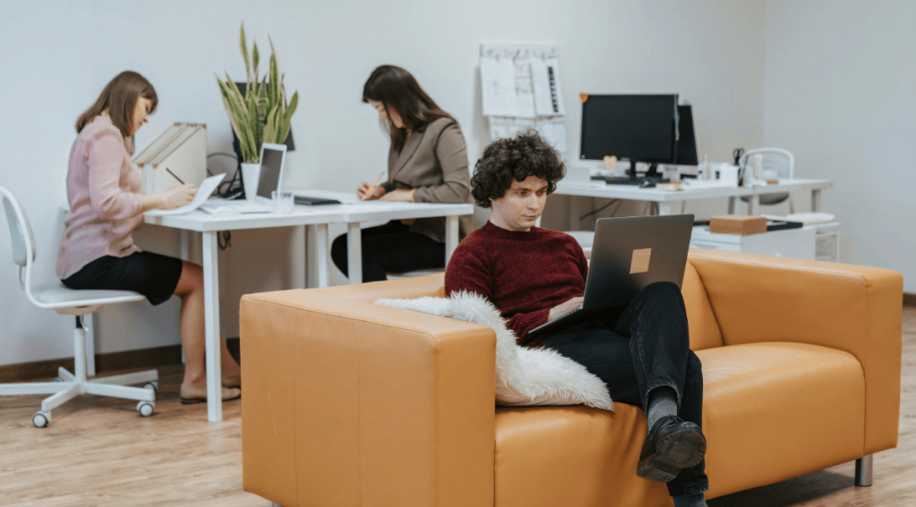 Persone al lavoro in ufficio moderno con postazioni flessibili, scrivanie condivise e area relax con divano, esempio di ambiente di lavoro efficiente.
